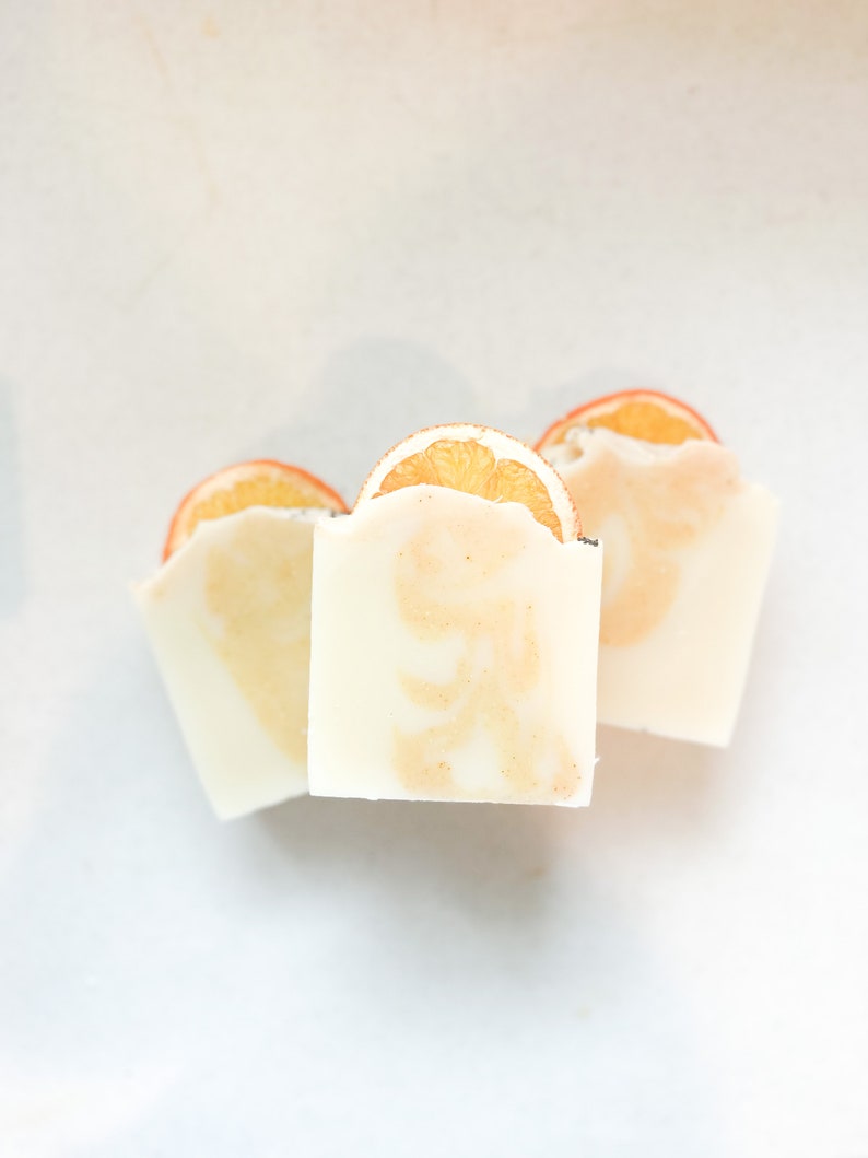 May include: Three white bars of soap with orange slices on top. The soap is a light white color with a subtle orange hue.