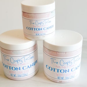 Op de afbeelding: Drie potten met geklopt suiker scrub met roze en witte inhoud. De etiketten zijn wit met blauwe tekst en zeggen "The Crafty Roses Bath, Body & Candle Co. Cotton Candy Whipped Sugar Scrub" en geven het nettogewicht in ounces en grammen aan.