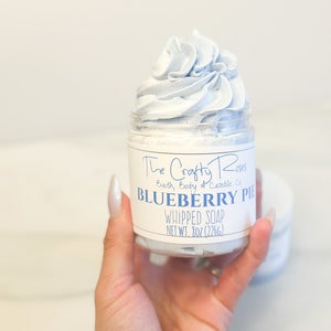 May include: A glass jar of light blue whipped soap with a white label that reads "The Crafty Roses Bath, Body & Candle Co. Blueberry Pie Whipped Soap Net Wt. 8oz (226g)"