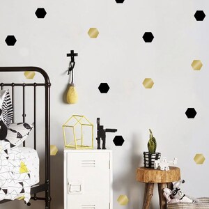 Peut inclure: Un mur blanc avec des autocollants hexagonaux noirs et dorés. Les autocollants sont disposés de manière aléatoire. Il y a un lit bébé, une table de chevet et une petite plante sur l'image.