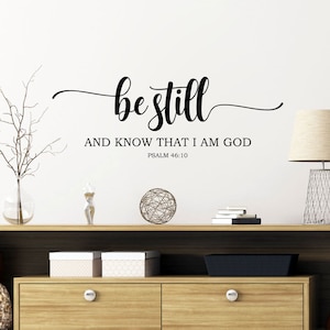Puede incluir: Pegatina de pared en blanco y negro con el texto "be still and know that I am God, Psalm 46:10". La pegatina está en una pared blanca sobre una estantería de madera con un jarrón, una esfera decorativa y libros.