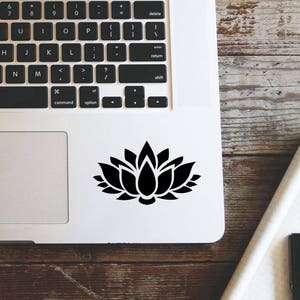 May include: A silver laptop computer with a black lotus flower sticker on the top right corner of the trackpad. The laptop is closed and sitting on a wooden surface.
