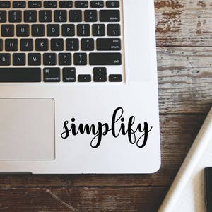 Puede incluir: Un portátil plateado con un teclado negro y la palabra "simplify" en escritura cursiva en la parte inferior derecha. El portátil está sobre una superficie de madera, con un vaso de líquido y auriculares blancos visibles.