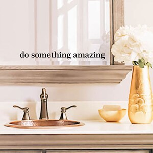 Puede incluir: Un tocador de baño con un espejo enmarcado, un lavabo de cobre y un jarrón dorado con flores blancas. El espejo tiene el texto "do something amazing" escrito en él.