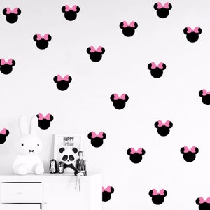 Puede incluir: Una pared blanca con calcomanías de la cabeza de Minnie Mouse en negro y rosa. Las calcomanías están dispuestas en un patrón aleatorio. Hay una figurita de conejo blanco y un marco de fotos con las palabras "Feliz cumpleaños" en una cómoda blanca.