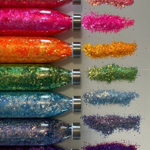 Könnte beinhalten: Eine Sammlung farbenfroher Glitzer in transparenten Flaschen mit silbernen Deckeln. Der Glitzer wird in einem Regenbogen von Farben präsentiert, darunter Rosa, Orange, Grün, Blau und Lila. Jede Flasche hat einen entsprechenden Glitzerhaufen daneben.
