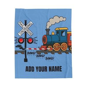 Può includere: Una coperta blu con un'illustrazione di un treno dei cartoni animati. Il design include un treno blu, un segnale di passaggio a livello e il testo "ADD YOUR NAME". Il treno è blu, rosso e giallo, con fumo.