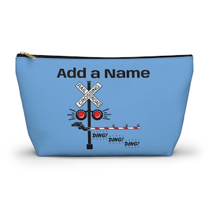 May include: A light blue zippered pouch with a railroad crossing graphic. The graphic includes a crossing sign, red lights, and a crossing gate. Text on the pouch reads "Add a Name" and "DING!" The pouch has a gold zipper.