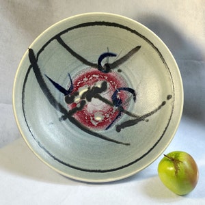 May include: A large, round ceramic bowl with a gray glaze and black and blue abstract designs. The bowl has a red and white center with black and blue accents.