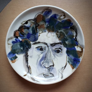 May include: A white ceramic plate with a hand-painted portrait of a person with blue and green hair. The person's face is painted in shades of blue, brown, and white.