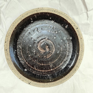 May include: A handmade ceramic plate with a dark brown, glossy interior and a speckled, light brown rim. The plate features a spiral design in the center and is suitable for serving food or as a decorative piece.