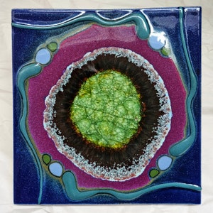 May include: A square ceramic tile with a blue background and a pink, white, and brown circle in the center. The circle has a green center with a cracked texture.