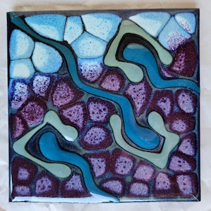 May include: A square ceramic tile with a colorful abstract design. The tile features a swirling pattern of teal, blue, green, and purple hues. The design resembles a flowing river or stream.