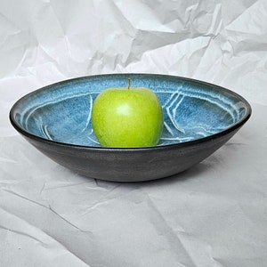 May include: A black ceramic bowl with a blue and white glaze pattern. A green apple sits in the center of the bowl.