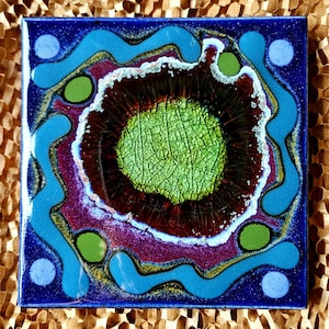 May include: Square ceramic tile with a vibrant abstract design. The tile features a dark blue border, with a central design of green, brown, purple, and blue. The design includes circular and wavy elements, creating a unique and colorful pattern.
