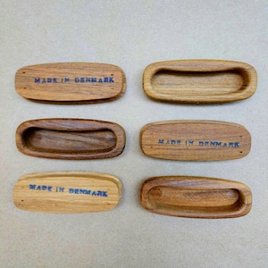 May include: Six wooden drawer pulls in various shades of brown. Each pull is rectangular with a curved handle recess. The text "MADE IN DENMARK" is printed in blue on the top of some of the pulls. The wood grain is visible.