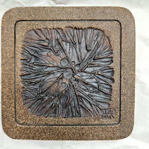 May include: A square, brown ceramic coaster with a textured, abstract design. The central design features a raised, spiky pattern with circular accents. The coaster has a raised border and appears handmade. The artist's signature is visible in the lower right corner.