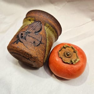May include: A small, brown ceramic pot with a carved butterfly design and a brown glaze. Next to the pot is a bright orange persimmon.