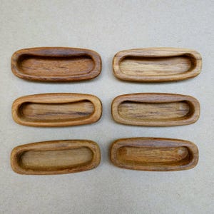 May include: Six wooden recessed drawer pulls. Each pull is oval-shaped with a curved handle area. The wood grain is visible, showing variations in color from light to dark brown. The pulls are arranged in two rows of three.