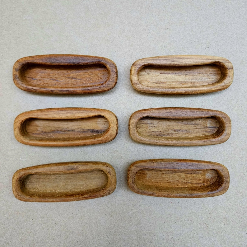 May include: Six wooden recessed drawer pulls. Each pull is oval-shaped with a curved handle area. The wood grain is visible, showing variations in color from light to dark brown. The pulls are arranged in two rows of three.