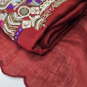 May include: A maroon fabric with intricate embroidery featuring geometric patterns, sequins, and beads. The fabric has a scalloped edge.