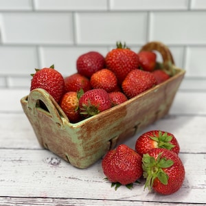 Può includere: Un cesto rettangolare in ceramica rustica pieno di fragole rosse mature. Il cesto ha una glassa verde e marrone, con un manico ad ogni estremità. Diverse fragole sono anche collocate fuori dal cesto su una superficie di legno bianco.