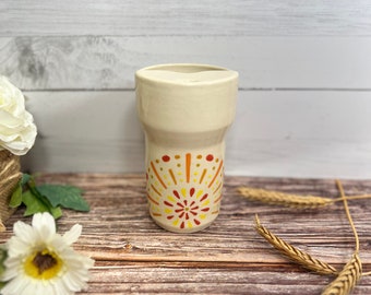 Op de afbeelding: Een witte keramische mok met een handgeschilderd ontwerp van een gele, oranje en rode zonnestralenpatroon. De mok staat op een houten oppervlak.