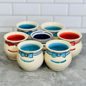 May include: Six ceramic candle holders with a white base and blue and red accents. Each holder features a blue bow and a cutout in the shape of a face. The holders are arranged in a circle on a gray surface.