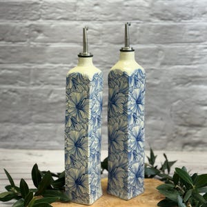 May include: Two matching ceramic oil bottles with stainless steel spouts. The bottles are rectangular with a cream-colored top and a blue floral pattern. They sit on a wooden cutting board with green leaves.