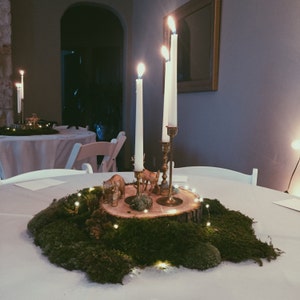 May include: A wooden slice centerpiece with moss, small white lights, and gold animal figurines. Three white candles are in gold candle holders.