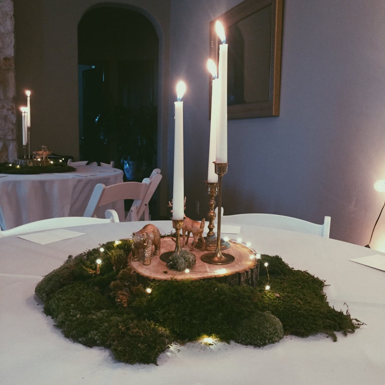 May include: A wooden slice centerpiece with moss, small white lights, and gold animal figurines. Three white candles are in gold candle holders.