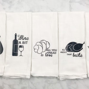 May include: Set of five white kitchen towels with black text and images. The towels feature sayings such as "Can't touch this", "Wine a bit", "All you knead is love", "All about that baste", and "Don't go bacon, my heart".