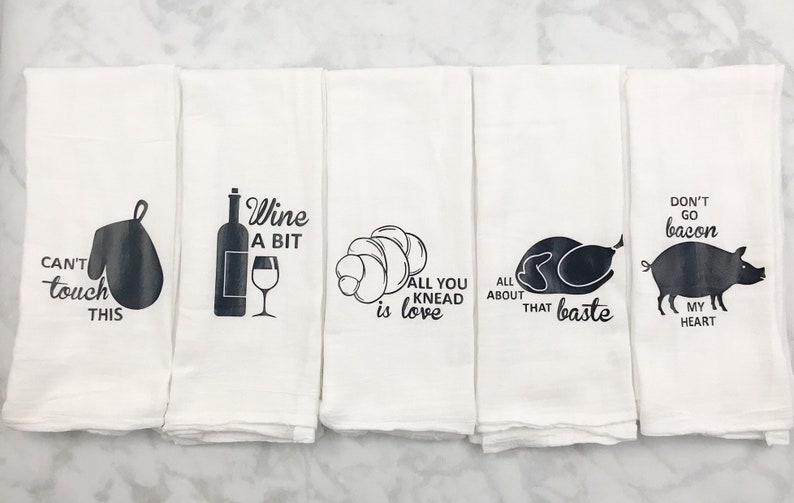 May include: Set of five white kitchen towels with black text and images. The towels feature sayings such as "Can't touch this", "Wine a bit", "All you knead is love", "All about that baste", and "Don't go bacon, my heart".