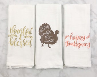 May include: Set of three white kitchen towels with Thanksgiving-themed designs. The first towel has gold text that reads "thankful & blessed". The second towel has a brown silhouette of a turkey with the text "gobble til you wobble". The third towel has orange text that reads "happy thanksgiving".