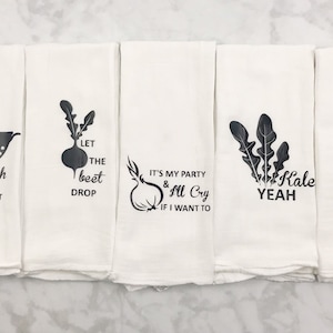 May include: Set of four white kitchen towels with black text and vegetable illustrations. The towels feature the phrases "wash rinse repeat", "let the beet drop", "it's my party & I'll cry if I want to", "kale yeah", and "olive me loves olive you".