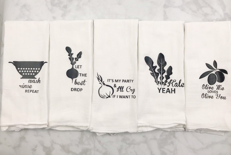 May include: Set of four white kitchen towels with black text and vegetable illustrations. The towels feature the phrases "wash rinse repeat", "let the beet drop", "it's my party & I'll cry if I want to", "kale yeah", and "olive me loves olive you".