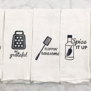 May include: Set of five white kitchen towels with black text and illustrations. The towels feature the following phrases: "Screw it", "Be grateful", "Flippin' awesome", "Spice it up", and "Shake it up".