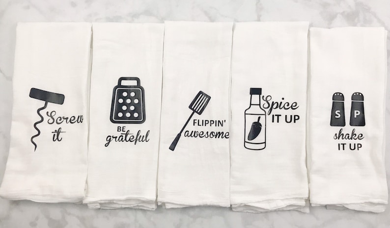 May include: Set of five white kitchen towels with black text and illustrations. The towels feature the following phrases: "Screw it", "Be grateful", "Flippin' awesome", "Spice it up", and "Shake it up".