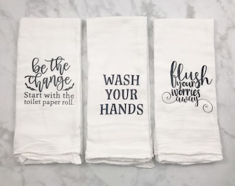 May include: Three white kitchen towels with black text. The first towel says "be the change start with the toilet paper roll." The second towel says "wash your hands." The third towel says "flush your worries away."