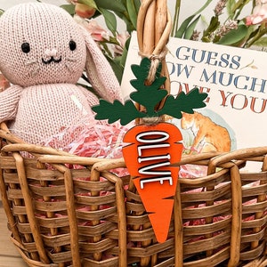 Puede incluir: Una cesta de mimbre llena de un conejito de peluche rosa, papel de seda rosa y un libro titulado "Adivina cu&aacute;nto te quiero". Una etiqueta de zanahoria de madera con el nombre "Olive" est&aacute; unida a la cesta.