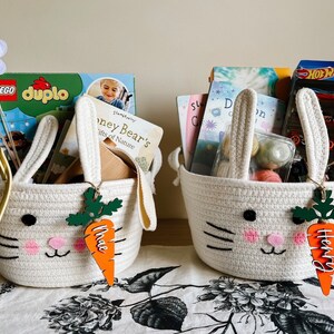 Puede incluir: Dos cestas blancas con forma de conejo llenas de libros y juguetes. Las cestas tienen etiquetas de madera con forma de zanahoria con los nombres "Mae" y "Henry" en ellas.