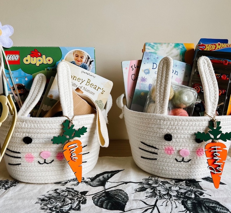 Puede incluir: Dos cestas blancas con forma de conejo llenas de libros y juguetes. Las cestas tienen etiquetas de madera con forma de zanahoria con los nombres "Mae" y "Henry" en ellas.