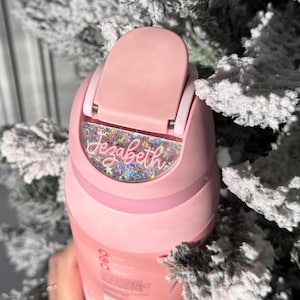 May include: A pink water bottle with a flip-top lid. The bottle features the name "Jezabeth" in a cursive font with glitter accents. The bottle is set against a backdrop of snow-covered evergreen branches.