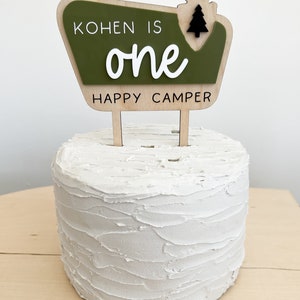 May include: A green and brown wooden cake topper with the text "Kohnen is one happy camper" and a small black pine tree graphic.