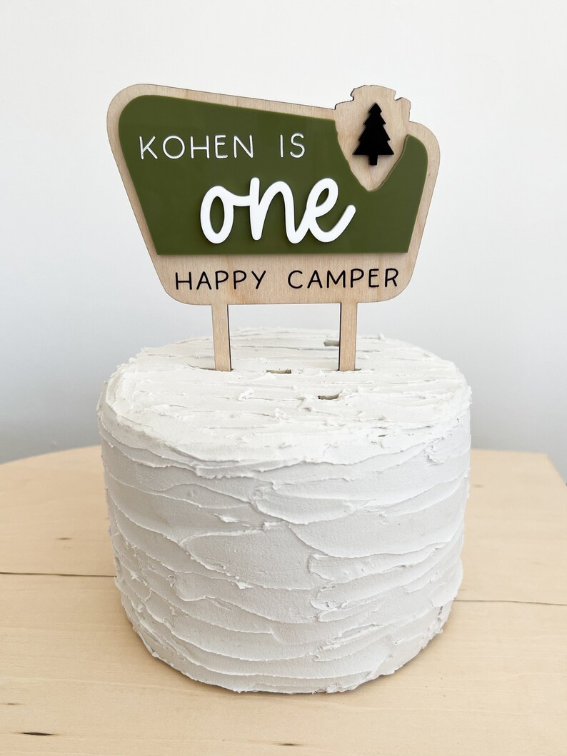 May include: A green and brown wooden cake topper with the text "Kohnen is one happy camper" and a small black pine tree graphic.