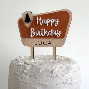 May include: A wooden cake topper with a brown and orange sign that says "Happy Birthday Luca" with a small black pine tree icon.