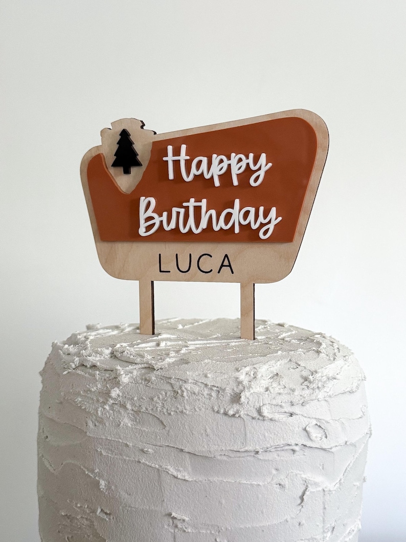 May include: A wooden cake topper with a brown and orange sign that says "Happy Birthday Luca" with a small black pine tree icon.