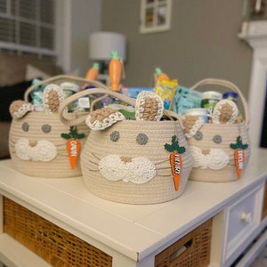 Puede incluir: Tres cestas de conejito beige llenas de golosinas de Pascua. Cada cesta tiene una etiqueta en forma de zanahoria con un nombre: Gavin, Henry y Declan.