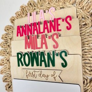 May include: A wooden sign with a white dry-erase surface and a banner that says "first day of". The sign has four separate sections with the names "Lila's", "Annalane's", "Mila's", and "Rowan's" written in pink, red, and green letters.