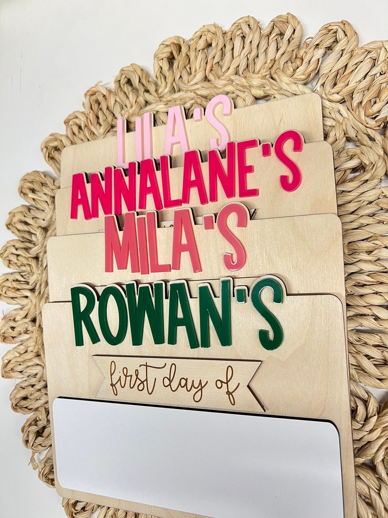 May include: A wooden sign with a white dry-erase surface and a banner that says "first day of". The sign has four separate sections with the names "Lila's", "Annalane's", "Mila's", and "Rowan's" written in pink, red, and green letters.
